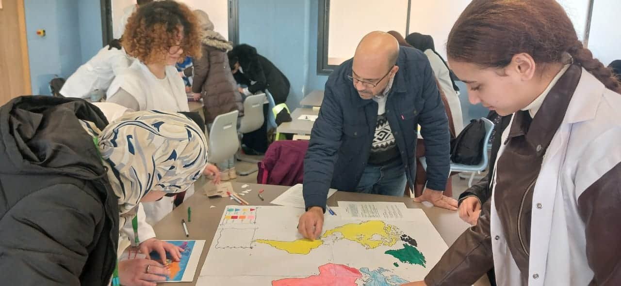 tep-workshop-facilitator-world-map-activity-morocco Facilitator and students working together around a large colorful world map during a TEP collective mapping exercise, with multiple groups visible in the background of a classroom in Morocco.